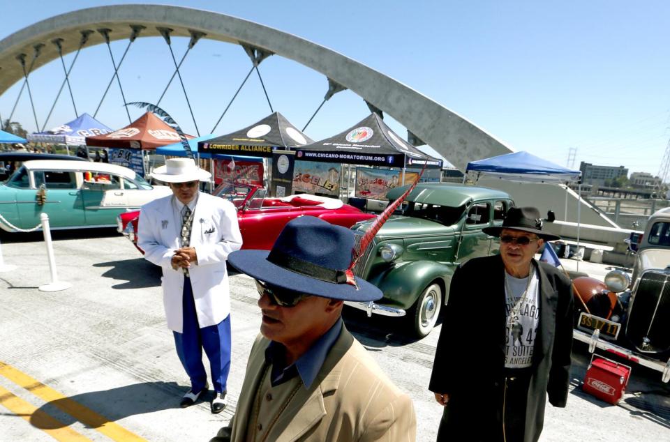 Men in long jackets, loose dress pants and hats with feathers stand outside vintage cars with a bring in the background.