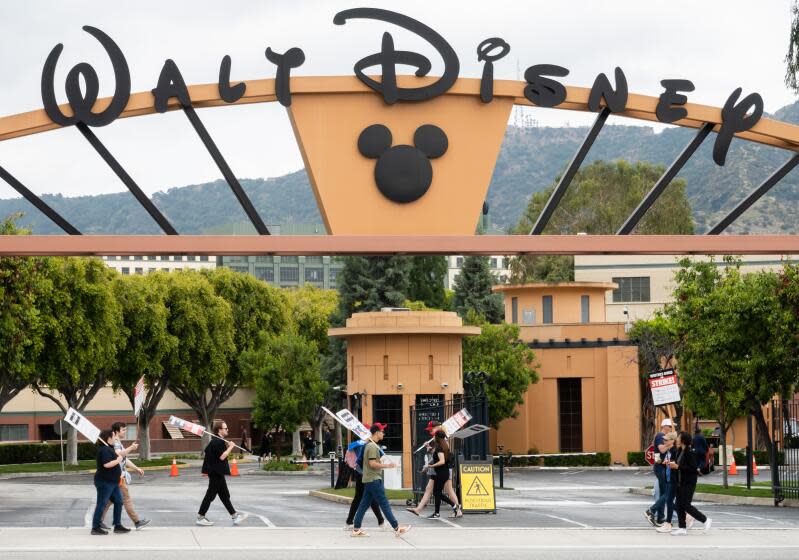 Picketers with signs walk on the sidewalk underneath the grand entrance to Walt Disney Studios.