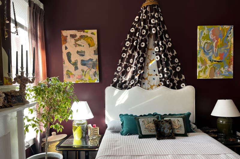 Marimekko fabric draped above white bed with white bedding in eggplant bedroom with colorful artwork and potted plant.