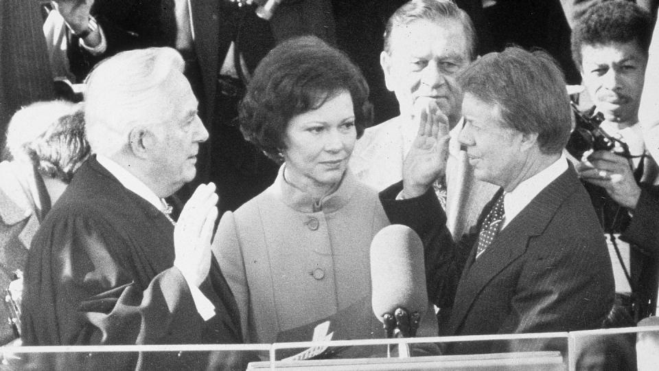 rosalynn carter stands and looks at jimmy carter whose right hand is raised as he faces earl burger whose right hand is also raised, in front of them is a podium with a large microphone, and several people are behind them
