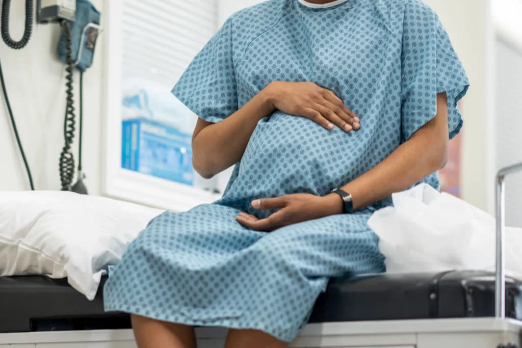 A pregnant woman of color sits on a medical examination table and cradles her midsection. She is dressed in a hospital gown.