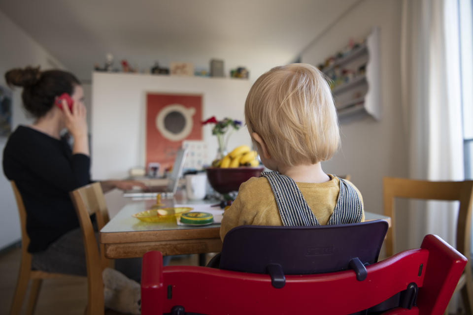 15.03.2020, Schweiz, Zürich: Eine Frau sitzt im Homeoffice an ihrem Laptop und telefoniert, während ihr Kind neben ihr in einem Kinderstuhl am Tisch sitzt. Die Schweiz hat das öffentliche Leben drastisch eingeschränkt, um die Verbreitung des neuartigen Coronavirus zu verlangsamen. Die Leute sind dazu angehalten, wenn möglich von zu Hause aus zu arbeiten. Foto: Christian Beutler/KEYSTONE/dpa +++ dpa-Bildfunk +++