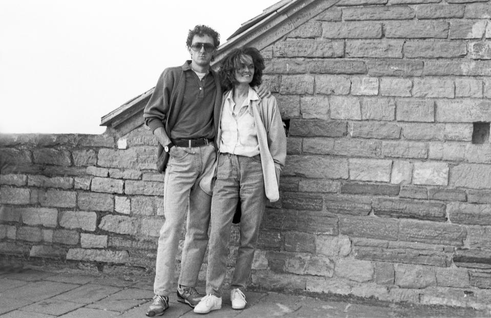 A couple stands together in front of a stone building. Both wear casual clothing with jeans and sunglasses