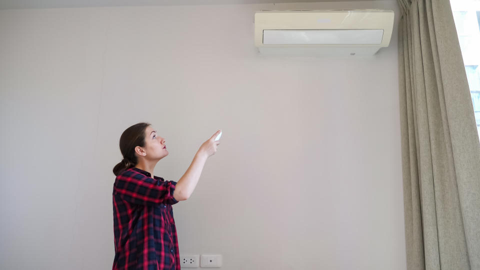 brunette with long hair in ponytail turns on air conditioner with remote control and smiles standing near white wall