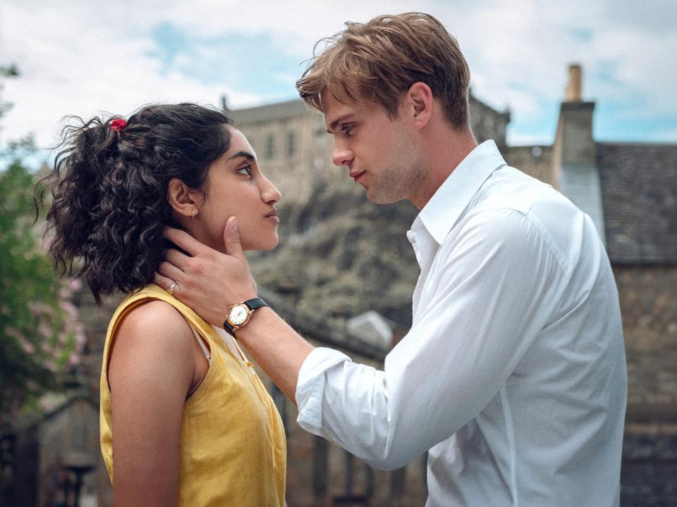 a young man cradles a woman's neck and jaw as they look intensely into each others' eyes face to face, standing outdoors in a picturesque village