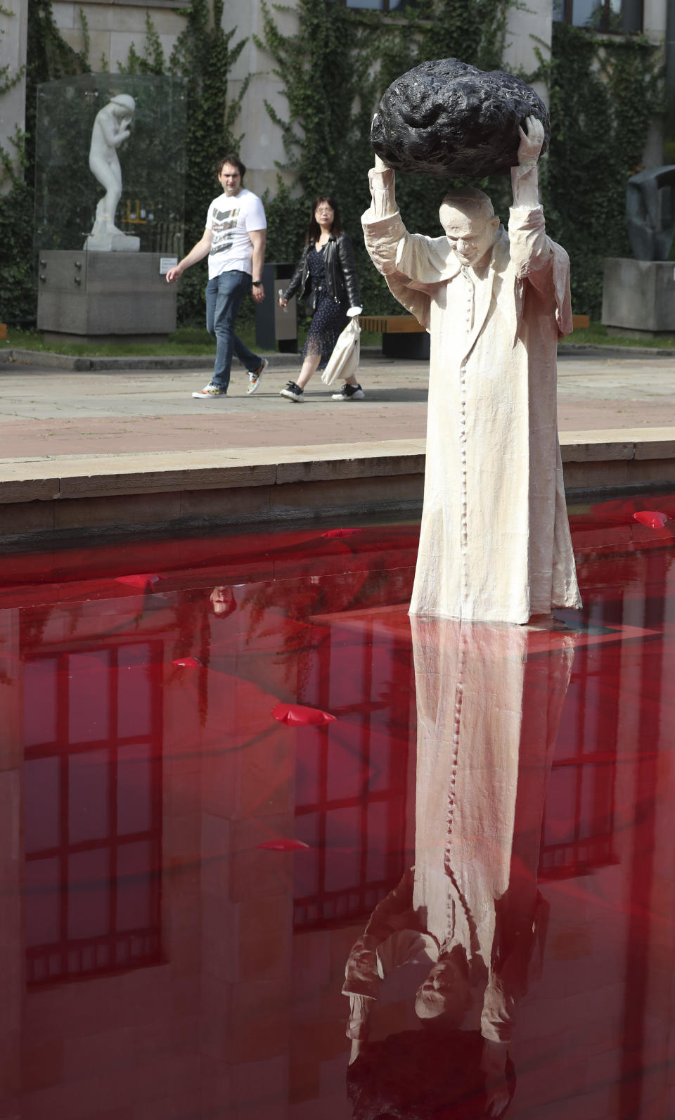 Visitors to the National Museum walk past a new statue of the late pope, St. John Paul II, throwing a stone at a "Poisoned Well" just hours before its official inauguration in the museum yard in Warsaw, Poland, Thursday, Sept. 24, 2020. The sculpture by Poland's Jerzy Kalina is said to be a response to a controversial 1999 sculpture by Italian Maurizio Cattelan in which the Polish-born pontiff was shown as being crushed by a similar stone. (AP Photo/Czarek Sokolowski)