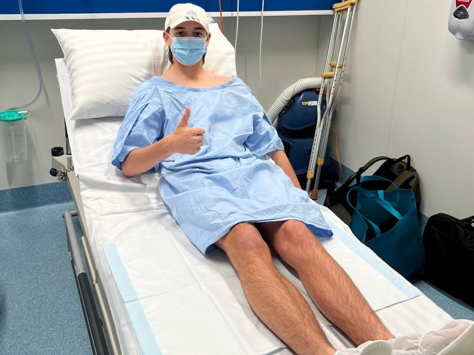 Image of Charlie sitting on a hospital bed wearing a surgerical mask, gown and surgical slippers. He wears a white baseball hat and gives a thumbs up to the camera with his right hand.