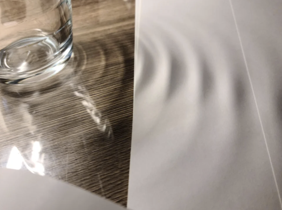 A glass of water is placed on a wooden surface, causing water ripples on a piece of paper next to it