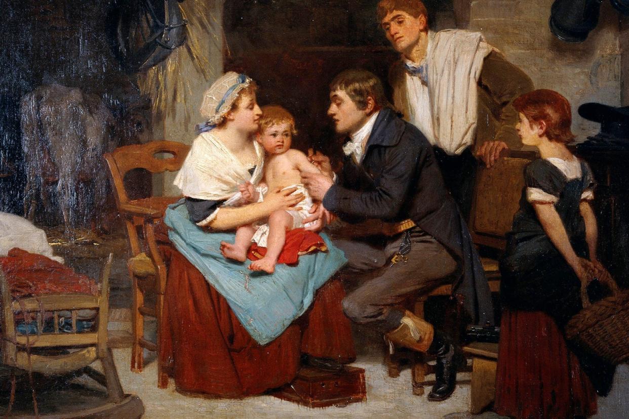 A woman holds a young child on her lap, while a doctor pricks the boy's arm. In the background, the boy's family members look on in curiosity.