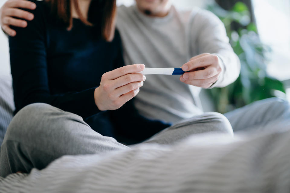 Two people sitting on a bed, holding a positive pregnancy test together. They are both wearing casual clothes