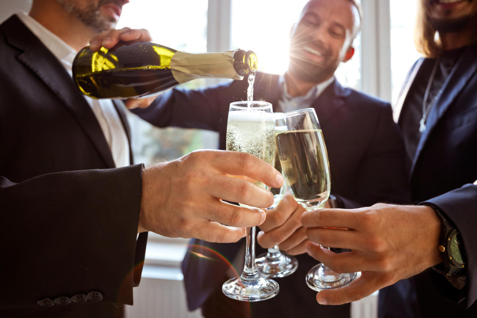 Three people in suits raising champagne glasses as one person pours a drink from a bottle, celebrating together.