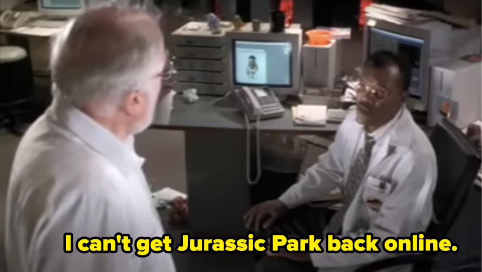 Two men in a laboratory setting, one seated at a desk with a computer showing a face, the other standing; Samuel L Jackson's character says he can't get Jurassic Park back online