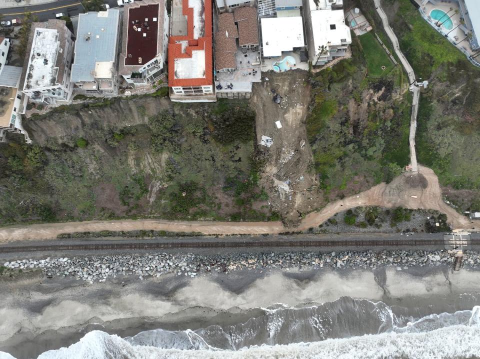 An overhead aerial view of homes on a cliff, a section where the land has given way, and ocean.