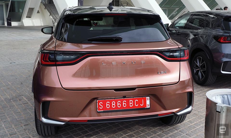 Bild eines Lexus LBX-Kofferraums, der unter dem Vordach der Stadt der Künste und Wissenschaften in Valencia geparkt ist.  Das Auto ist in „Sonic Copper“ lackiert, einer Art metallischem Orange.
