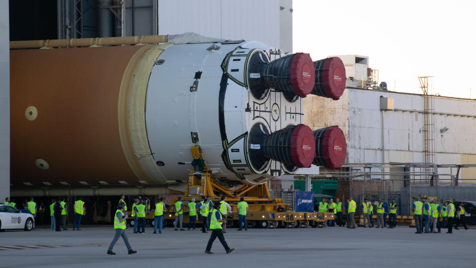  Fitted red tarps wrap around the circular bell ends of four humungous rocket engines embeded in a white boattail mount on an orange fuel tank, flipped horizontal, with the bulk of its fuselage hiddenn behind the doors or a large hangar. Workers in highlighter vests scurry about like ants. 