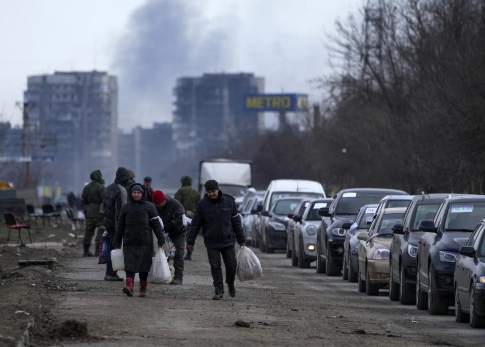 People walk away from a smoking city alongside a line of waiting cars.