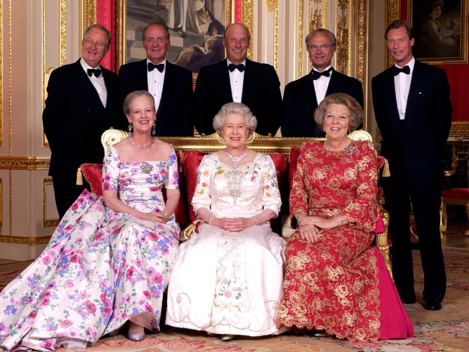 The Queen sitting and smiling in a white satin ballgown covered in pastel floral embroidery down the center and on the chest and three-quarter sleeves. She's wearing a large diamond necklace. She's sitting next to two women in colorful floral gowns and five men in tuxedos stand behind them.