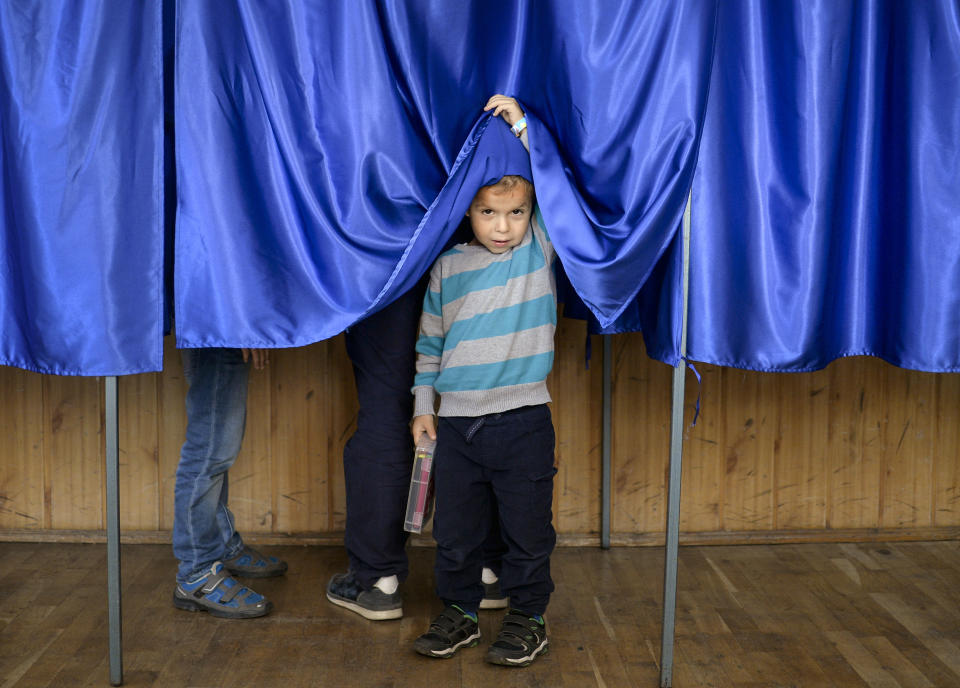 A child lifts the courtain of a voting booth as his father casts his vote, in Ciorogarla, Romania, Sunday, Oct. 7, 2018. Romanians are voting Sunday around the country for a second day on a constitutional amendment that would make it harder to legalize same-sex marriage. (AP Photo/Andreea Alexandru)