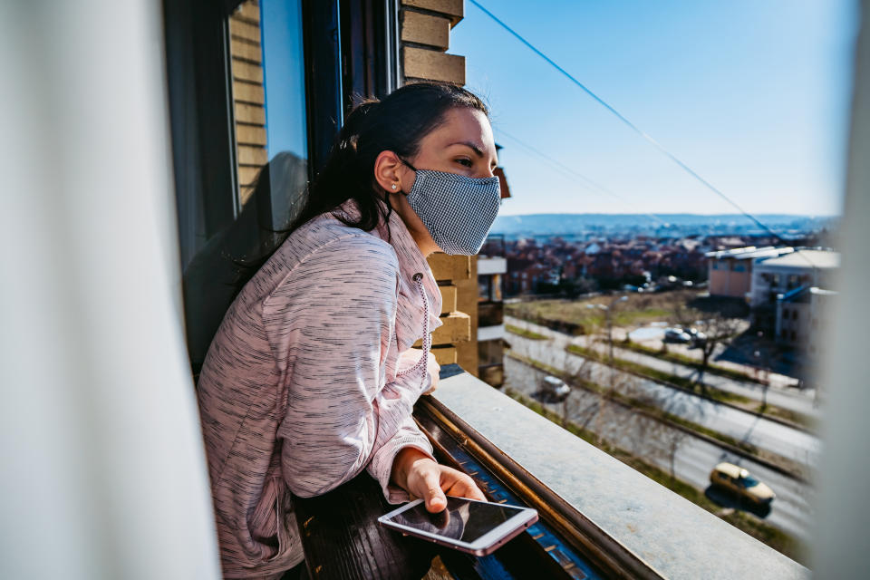 A woman looks out of a city window. She's wearing a cloth face mask.