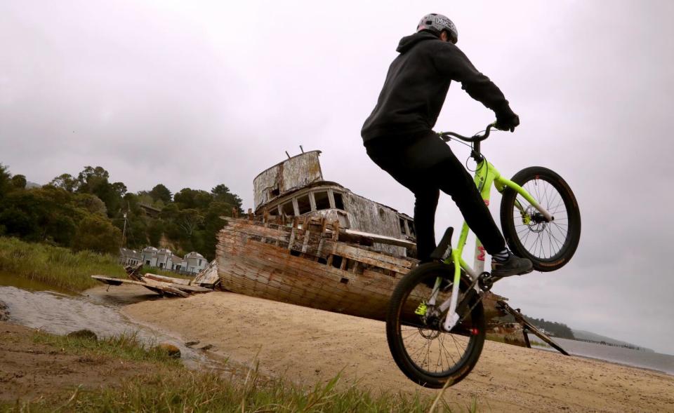 A young man riding a yellow bike hops over a creek near a listing boat.