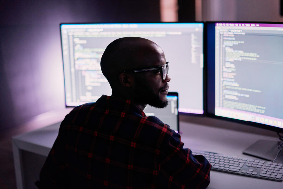 Man in glasses working at a computer with code on screens, in a dimly lit room, suggesting a programming or tech environment