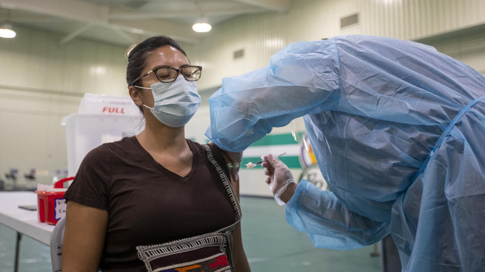A health worker wearing rubber gloves and full coveralls gives a shot into the upper arm of a seated woman who is wearing a face mask and has her eyes closed.