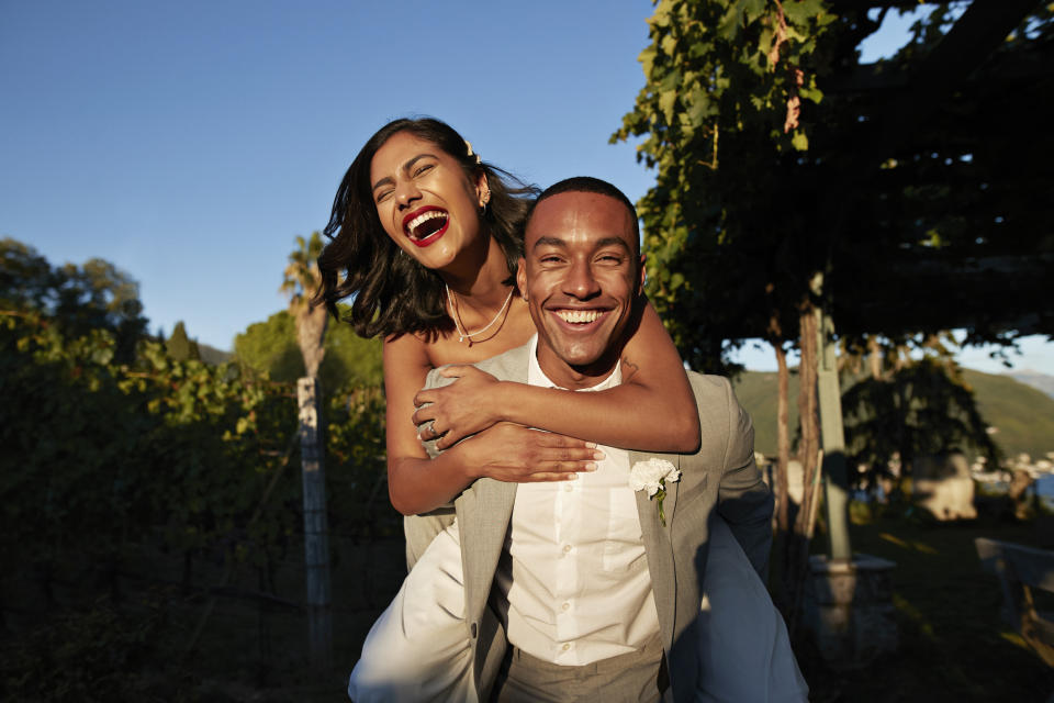 A woman with red lipstick in a white dress joyfully piggybacks on a man in a gray suit, both smiling broadly at their wedding