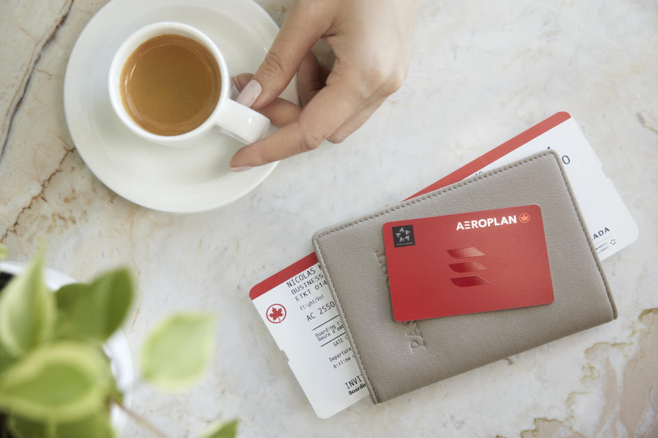Travel concept, a woman's hand on an espresso cup next to an Aeroplan credit card, passport wallet and Air Canada plane ticket