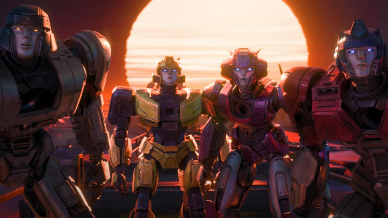  Image from the animated movie 'Transformers One.' Four transformers are standing in a row looking forwards. There is a large sun in the background. 