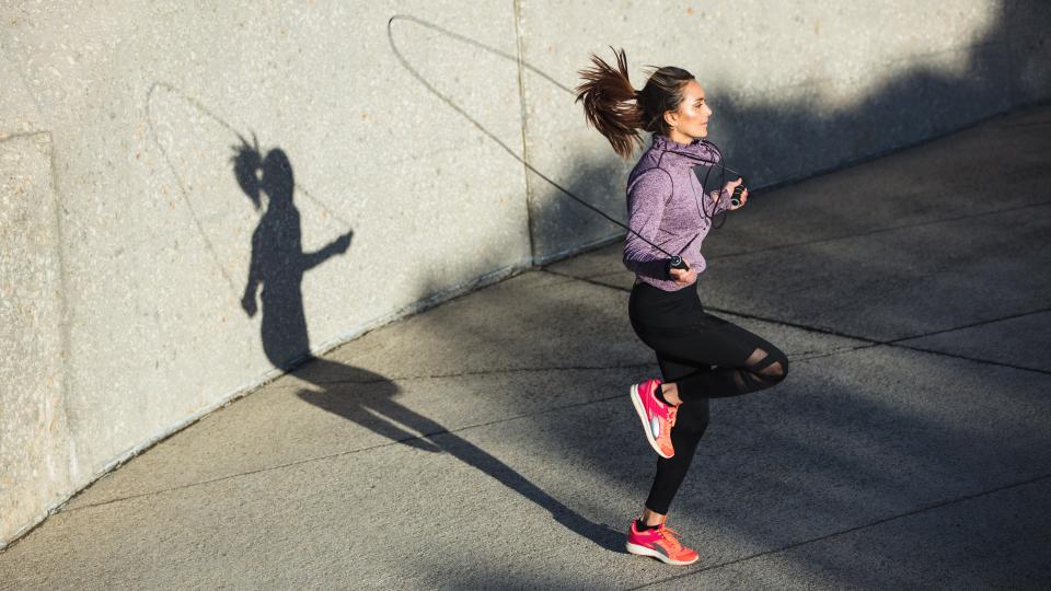 Woman jumping rope with right knee bent while jumping outdoors against a wall backdrop