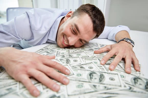 A smiling man resting his head and hands on a pile of cash on his desk.
