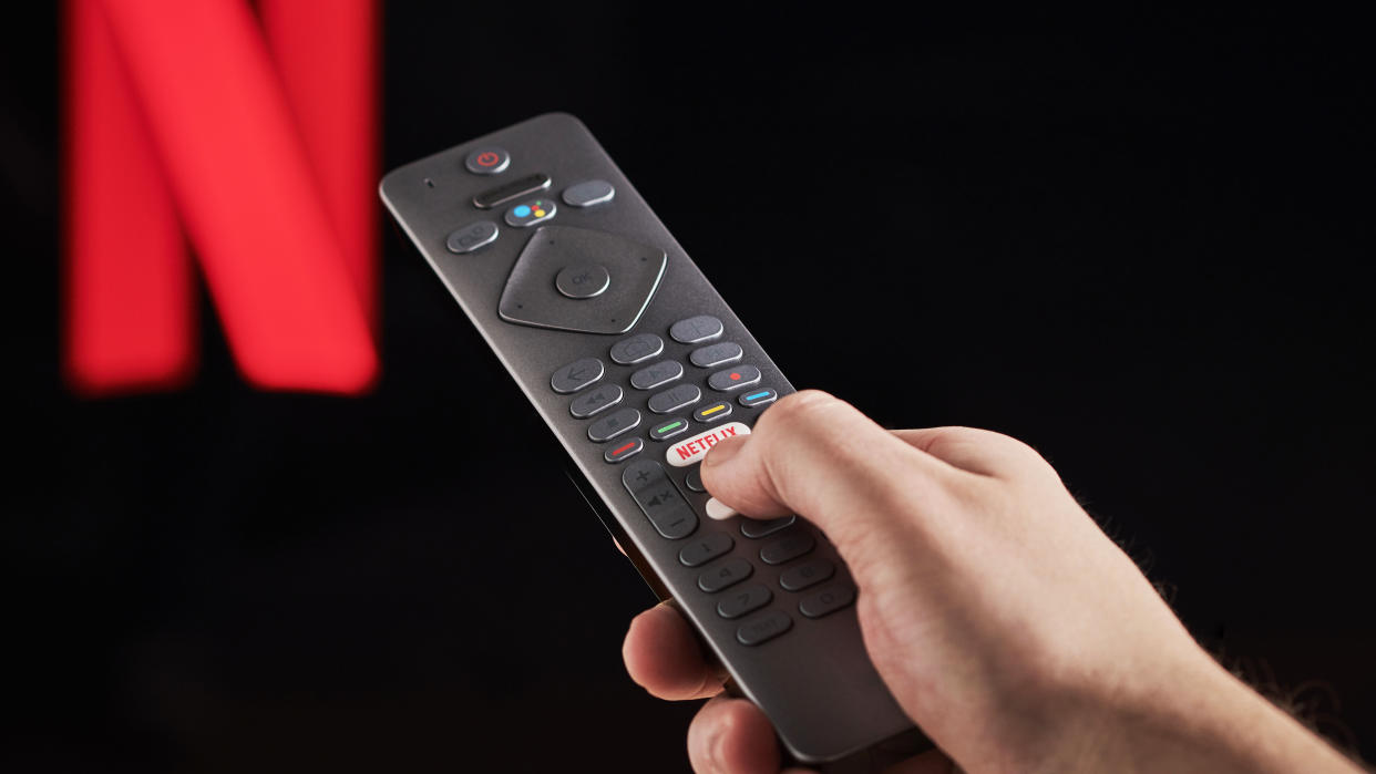  An image of a person holding a TV remote and pressing the Netflix button. In the background is a black screen with the Netflix logo. 