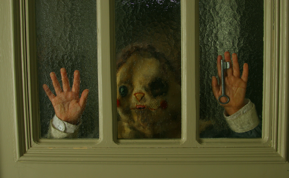 A child with a cloth clown-like face holding his hands against the door and a key in his hands