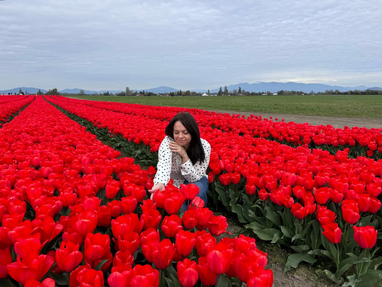 Bernadette, wearing a polka dot long-sleeve shirt, squats down in a field of red tulips and touches a flower.