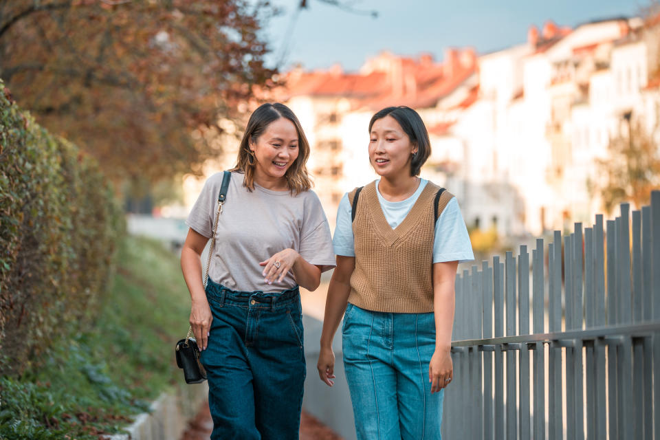 Two Asian female best friends talking and laughing while walking home from school along a river in old town, wearing fashionable clothes and relaxed with afternoon light in the background.