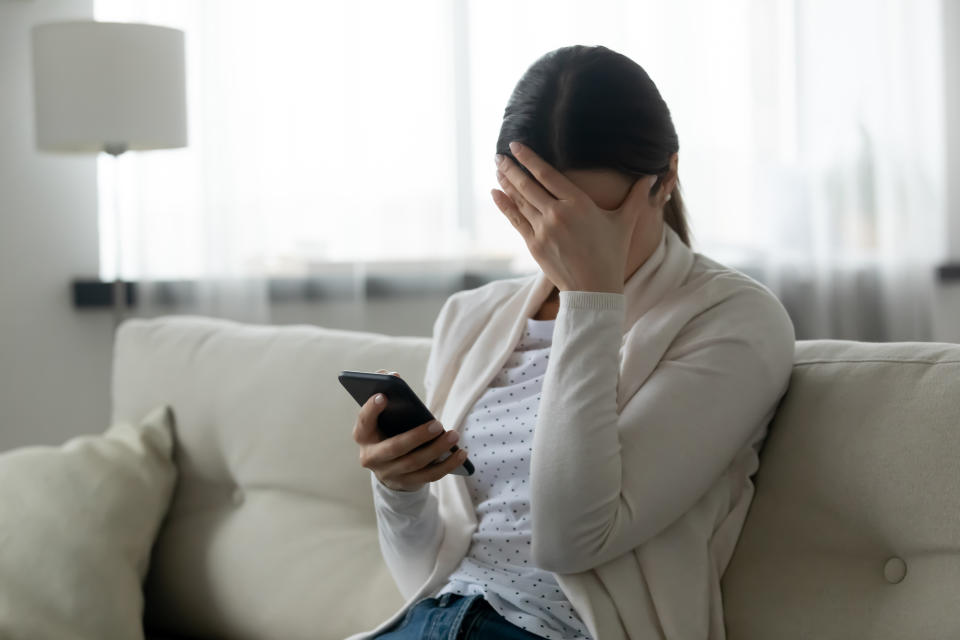 Woman sit on sofa holding smartphone cover face with hand feels scared humiliated suffering from cyberbullying being on-line abused by stalker. Bad news, life troubles, break up with boyfriend concept