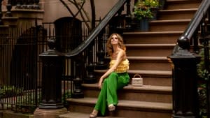 Red-haired young woman in stylish bright yellow top and green trousers elegant sandals sitting on the potted flowers porch stairs on Manhattan West village.