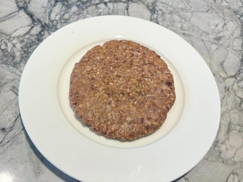 A grayish raw beef burger patty on a white plate placed on a gray table