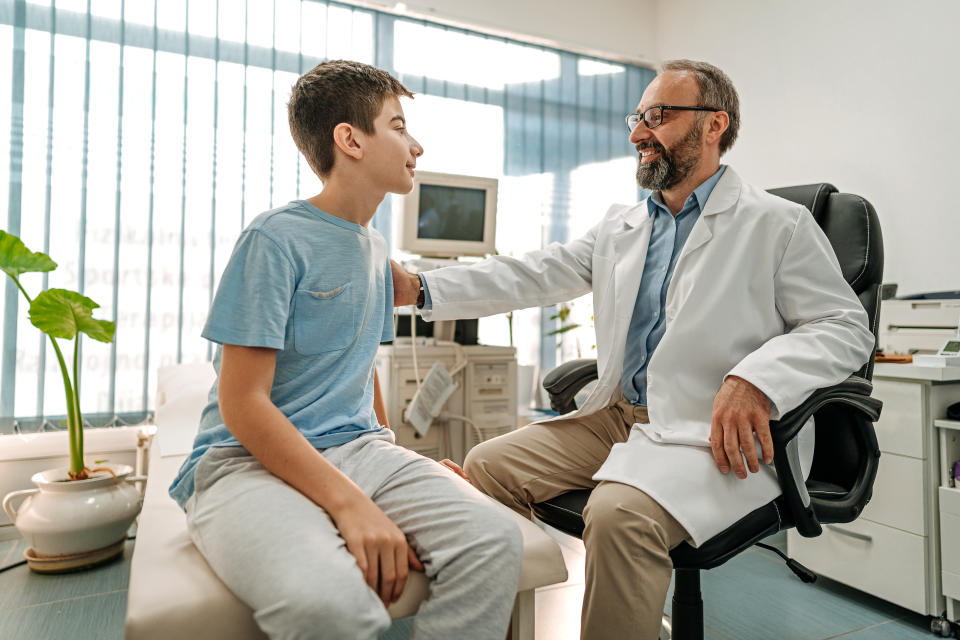 A doctor in a white coat sits in an office chair, smiling and talking with a young boy seated on an examination table