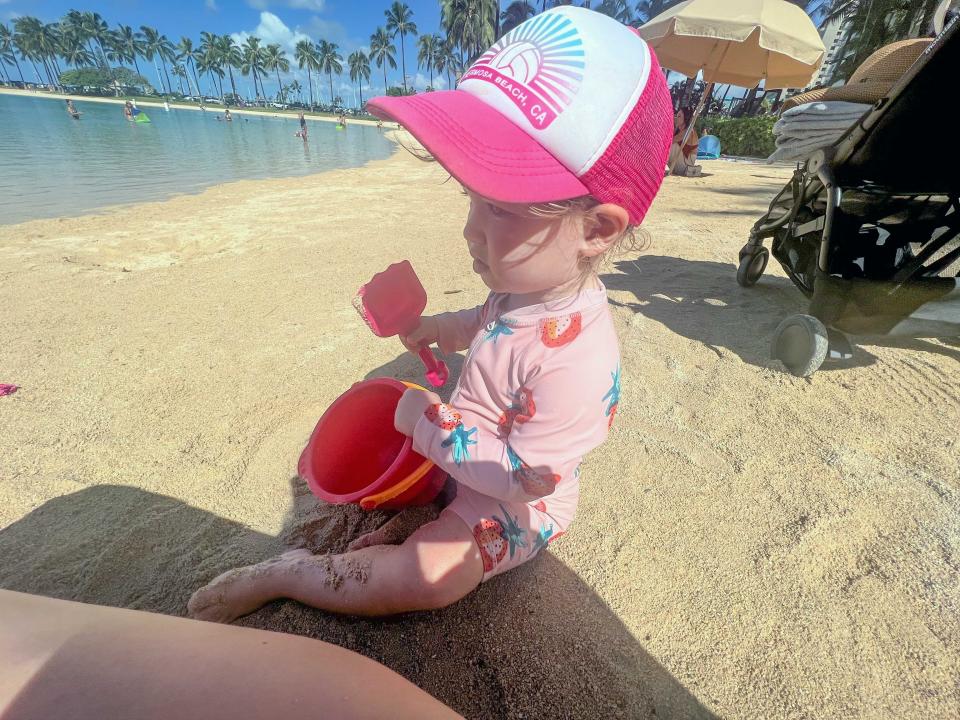A child on the beach wearing a bathing suit and hat and holding a pail and shovel.
