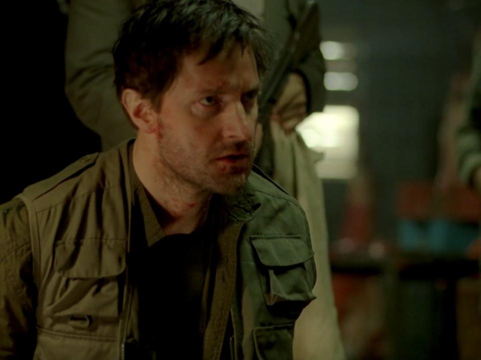 richard armitage as john porter, crouching on the ground with his hands restrained as someone holds a gun behind him