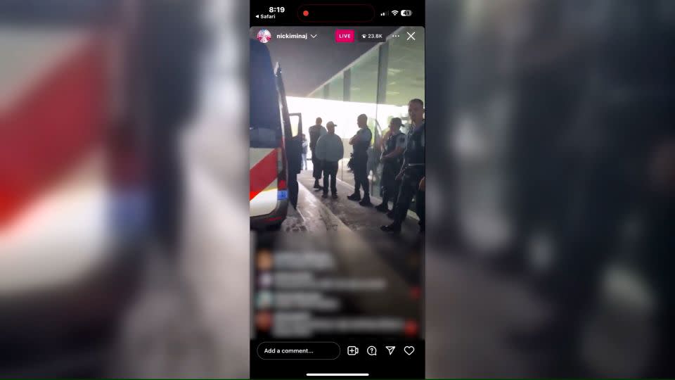 In this still from Nicki Minaj's Instagram Live, the rapper is ushered out of her vehicle by police at Amsterdam’s Schiphol Airport. CNN has blurred the comments on the livestream. - From Nicki Minaj/Instagram