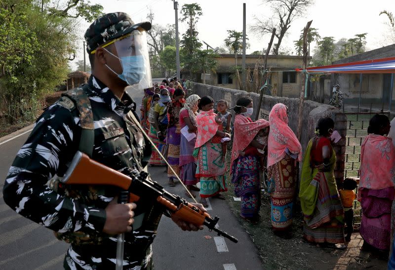 An armed policeman wearing a face shield stands guard as women wait in line to cast their votes outside a polling booth during the first phase of the West Bengal state election in Purulia