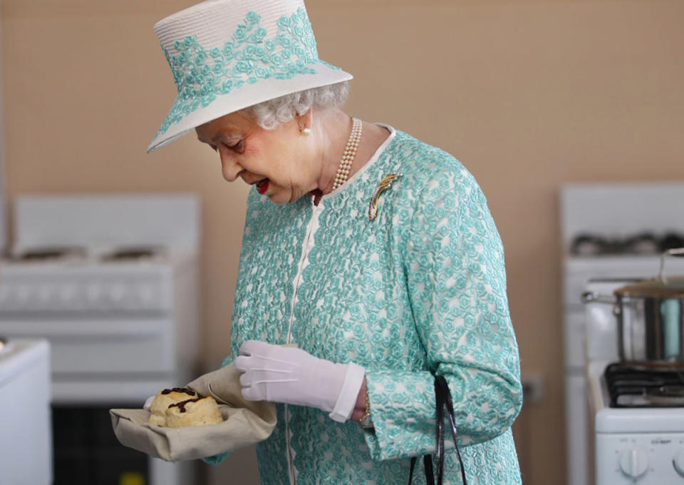 The Queen holds two scones while wearing white gloves and a white hat with blue detail on it