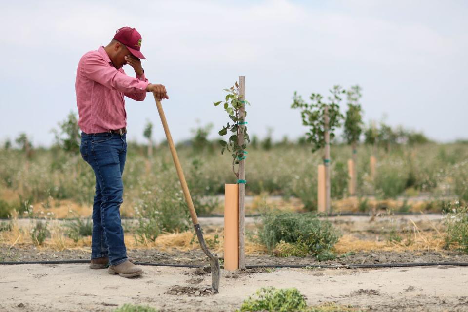 A man wearing a red baseball cap holds a shovel in one hand and wipes his brow with the other.