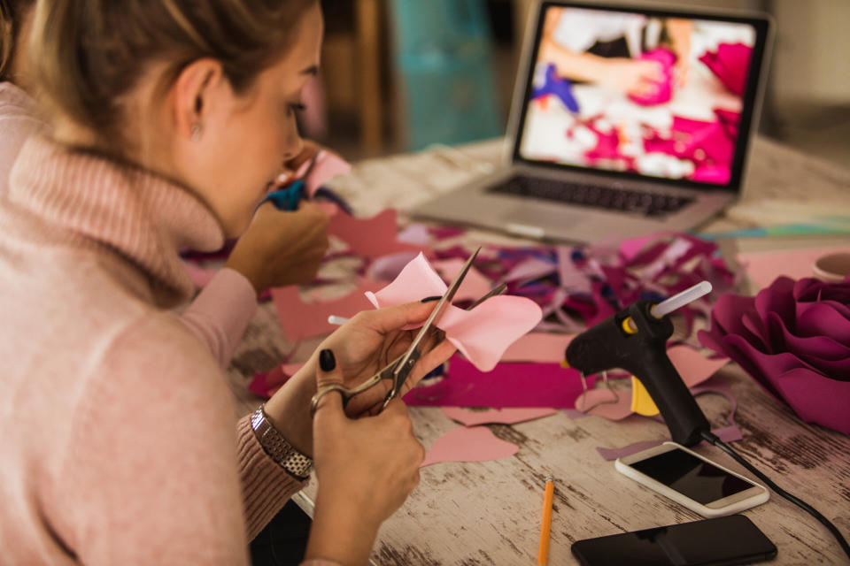 Two women crafting with scissors, paper, and a glue gun at a table, with a laptop showing a crafting tutorial in the background