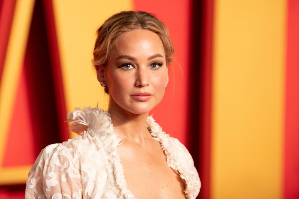 Jennifer Lawrence on the red carpet, wearing a dress with feathered details and minimal jewelry, poses in front of a colorful backdrop