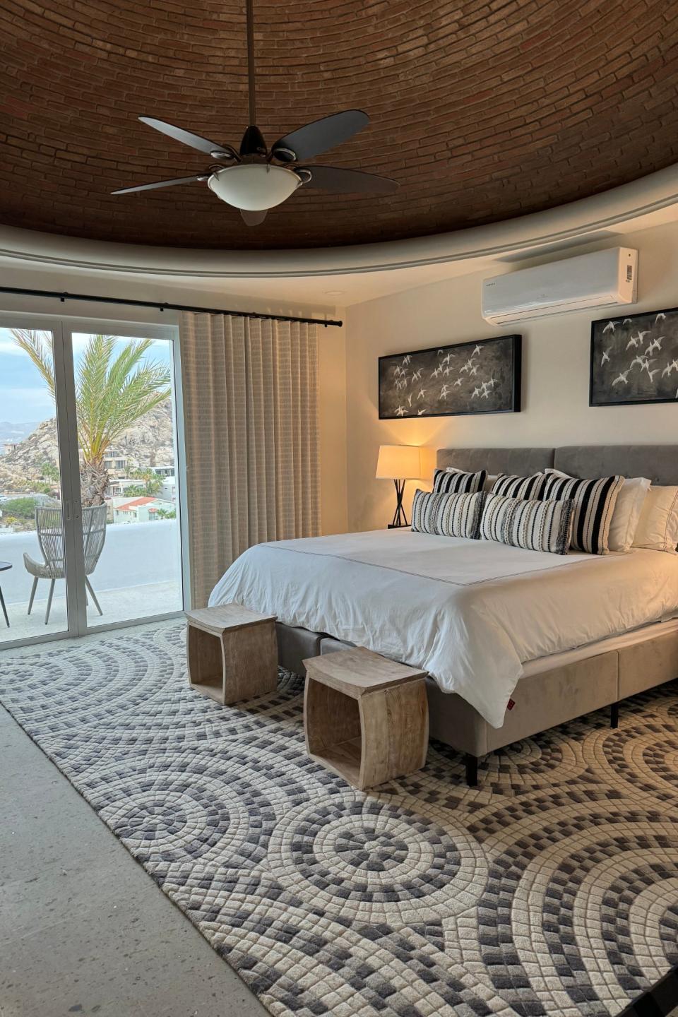 A modern and spacious bedroom with a circular ceiling, a large bed with striped pillows, two wooden stools at the foot, and a view of outdoor scenery through a sliding door