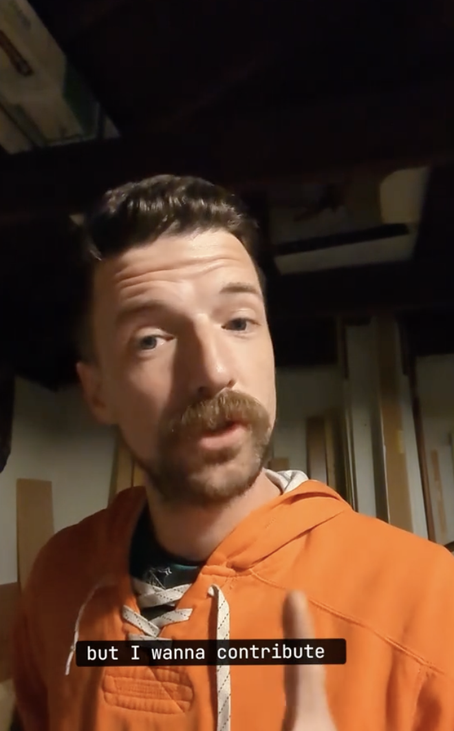 Man with mustache in orange jacket, speaking, subtitled 