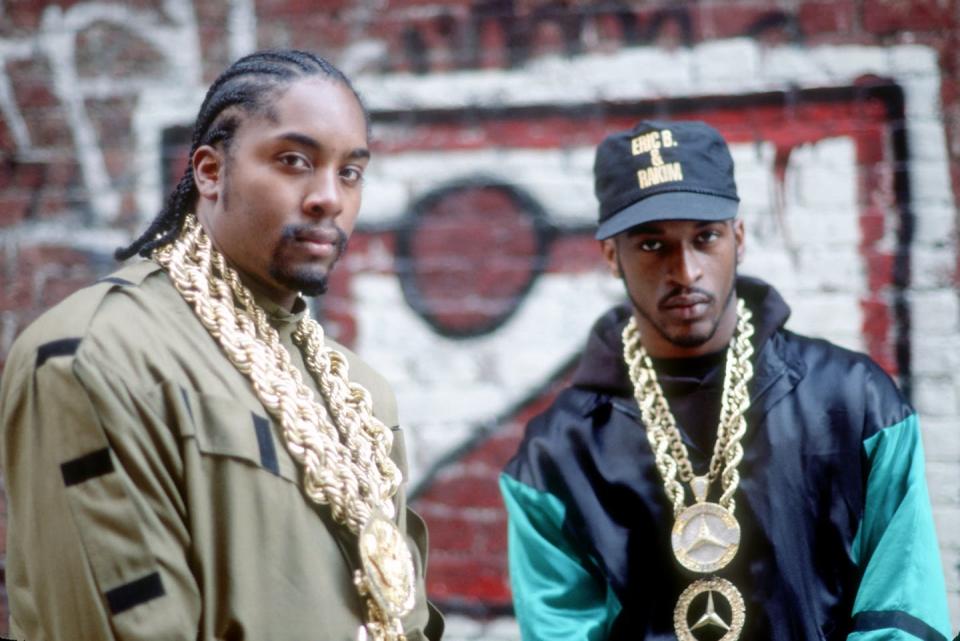 Two rappers wearing golden chains pose for a photo in front of a wall covered in graffiti.